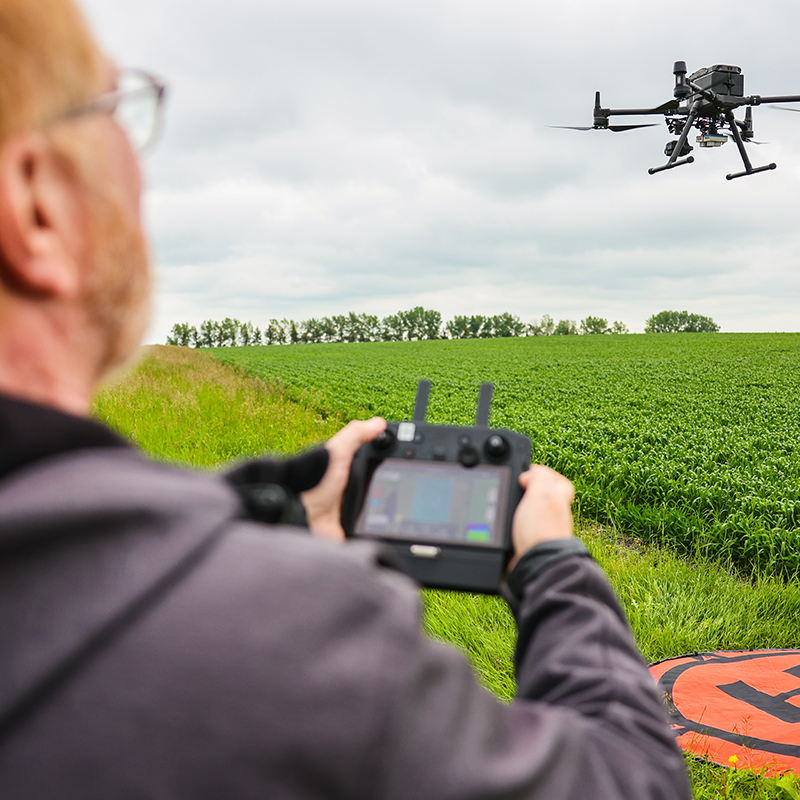 A person operating a drone controller in the foreground while a large drone hovers over a green agricultural field. A landing pad is visible on the ground, and the sky is overcast, indicating a possible crop survey or monitoring activity.